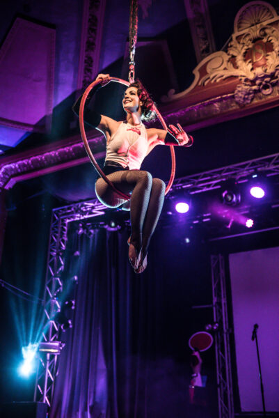 Aerial burlesque artist suspended from Komedia's ceiling in an acrobatic hoop, posing above the audience below