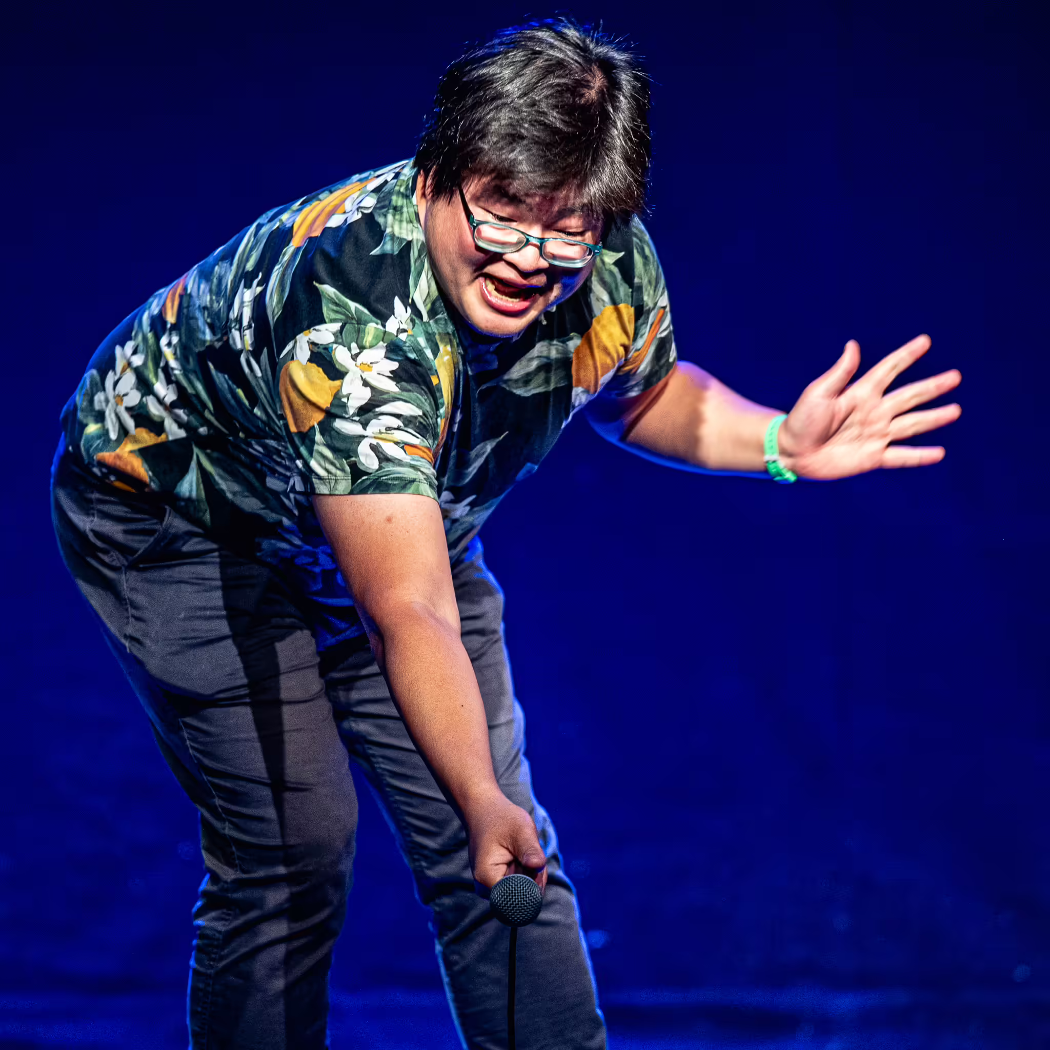 A comedian wearing a bright Hawaiian shirt, pointing their microphone into the audience.